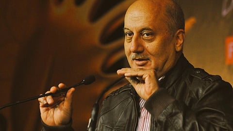 माइक के सामने खड़े होकर बोलते अभिनेता अनुपम खेर जिन्होंने काले रंग की जैकेट पहनी है 
