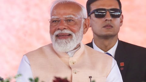 भारत के प्रधानमंत्री नरेंद्र मोदी गुलाबी रंग के जैकट और सफ़ेद शर्ट पहने हुए हैं और सामने देखकर मुस्कुरा रहे हैं। पीछे काले कपड़ों में उनके सुरक्षा कर्मी दिख रहे हैं। 
