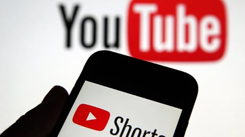 एक व्यक्ति ने हाथ में मोबाइल पकड़ा है जिसपर शॉर्ट्स लिखा है जबकि पीछे बैकग्राउंड पर यूट्यूब लिखा दिख रहा है। 
