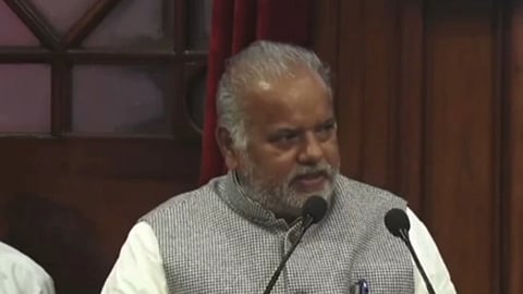 मंच पर माइक्रोफोन के सामने खड़े एक वरिष्ठ व्यक्ति सभा को संबोधित करते हुए बोलते दिखाई दे रहे हैं।