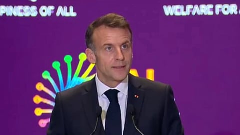 काले रंग का सूट पहने हुए फ़्रांस के राष्ट्रपति मैक्रों, माइक के सामने खड़े होकर कुछ बोलते हुए 
