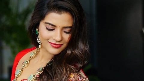 अभिनेत्री साड़ी पहने हुई है और पोज दे रही है। पीछे कुछ पौधे दिखाई दे रहे हैं। 
