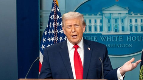 अमेरिका के राष्ट्रपति डोनाल्ड ट्रंप माइक के सामने खड़े होकर बोलते हुए और उन्होंने नीला कोट, लाल टाई और सफ़ेद शर्ट पहना है। 
