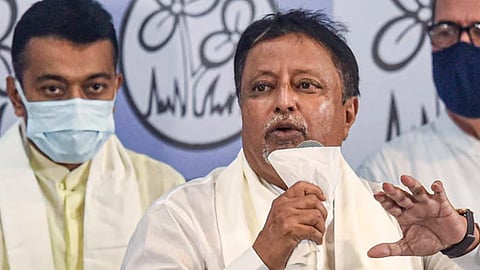 मंच पर माइक्रोफोन पकड़े भाषण देते हुए वरिष्ठ नेता मुकुल रॉय, पीछे पार्टी का चिन्ह और सहयोगी दिखाई दे रहे हैं।
