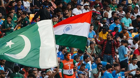 स्टेडियम में भारत और पाकिस्तान के क्रिकेट प्रशंसक साथ बैठे हैं, एक व्यक्ति भारतीय तिरंगा लहरा रहा है जबकि दूसरे दर्शक पाकिस्तानी झंडा पकड़े हुए मैच का आनंद ले रहे हैं।