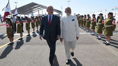 इज़राइल में औपचारिक गार्ड ऑफ ऑनर के दौरान प्रधानमंत्री नरेंद्र मोदी और इज़राइली प्रधानमंत्री बेंजामिन नेतन्याहू साथ चलते हुए दिखाई दे रहे हैं, दोनों ओर सैनिक पंक्तिबद्ध खड़े हैं।