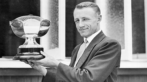 हाथ में ट्रॉफी लिए एक क्रिकेटर जिसने सूट पहन रखा है 
