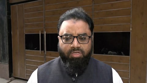 चश्मा और काली दाढ़ी वाले एक व्यक्ति घर के अंदर कैमरे के सामने बोलते हुए नजर आ रहे हैं।