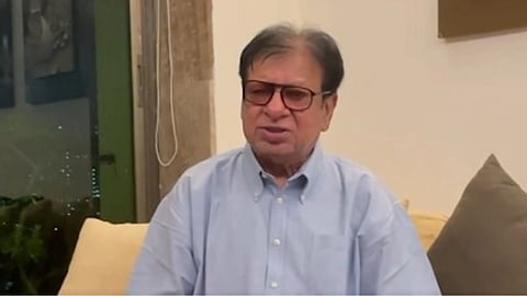 सोफे पर एक व्यक्ति बैठा है, जिसने नीले रंग की शर्ट पहनी है और चश्मा लगाया हुआ है 
