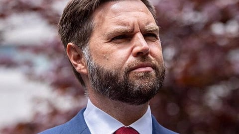 अमेरिका के उपराष्ट्रपति जेडी वेंस का क्लोज़-अप चित्र, नीले सूट और लाल टाई में, बाहर खड़े हुए गंभीर भाव के साथ, पृष्ठभूमि में धुंधले पेड़ दिखाई दे रहे हैं।