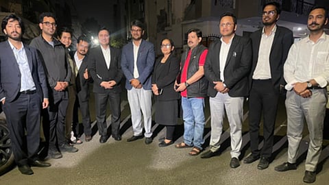 रात के समय एक आवासीय परिसर के बाहर औपचारिक कपड़ों में खड़े युवाओं और पेशेवरों का समूह फोटो के लिए पोज़ देता हुआ दिखाई दे रहा है।