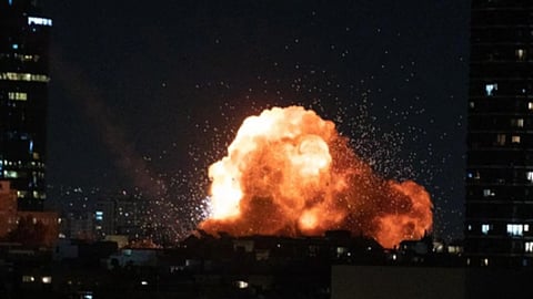 रात के समय शहर की इमारतों के बीच तेज धमाके के साथ उठता हुआ विशाल आग का गोला और धुएं का गुबार दिखाई दे रहा है।