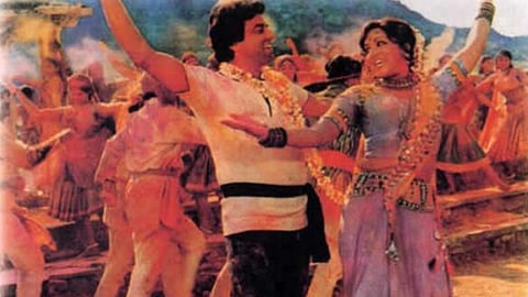 एक फिल्मी दृश्य में पुरुष और महिला रंगीन कपड़ों में नाचते हुए दिखाई दे रहे हैं, पीछे कई लोग भी जश्न मना रहे हैं।