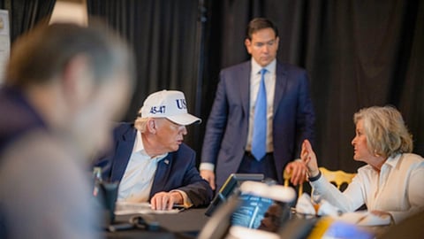 डोनाल्ड ट्रंप सफेद टोपी पहने एक कॉन्फ्रेंस टेबल पर अधिकारियों के साथ चर्चा करते हुए बैठे हैं, जबकि एक अन्य व्यक्ति उनके पीछे खड़ा है और कमरे में गहरे रंग के पर्दे लगे हैं।