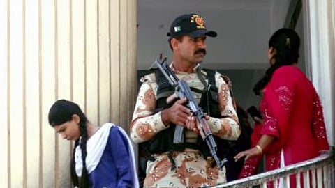 पाकिस्तान में सुरक्षा बल का जवान बंदूक के साथ खड़ा, पास से गुजरते छात्र-छात्राएं।