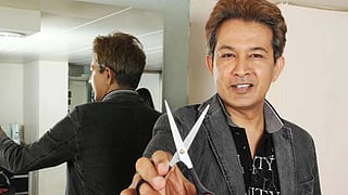 जावेद हबीब की एक क्लोज-अप तस्वीर, जिसमें उन्होंने ग्रे रंग का ब्लेज़र पहना हुआ है और हाथ में कैंची थामी हुई है। उनके पीछे एक शीशा है जिसमें उनकी पीठ दिख रही है।
