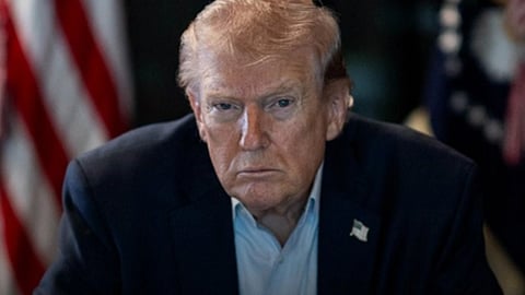 नीले रंग की सूट में राष्ट्रपति ट्रंप, गंभीरता से देखते हुए और पीछे अमेरिका का झंडा 
