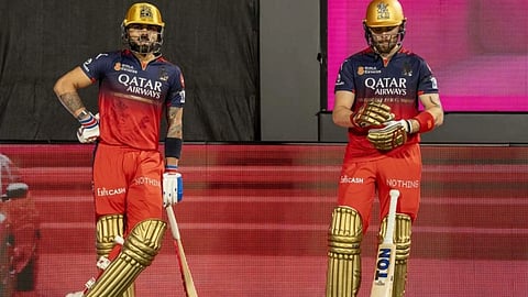 रॉयल चैलेंजर्स बेंगलुरु (RCB) की जर्सी, सुनहरे हेलमेट और पैड्स पहने बल्लेबाज मैदान पर बल्लेबाजी के लिए तैयार खड़े हैं। दोनों ने हाथ में बल्ला पकड़ा हुआ है। 
