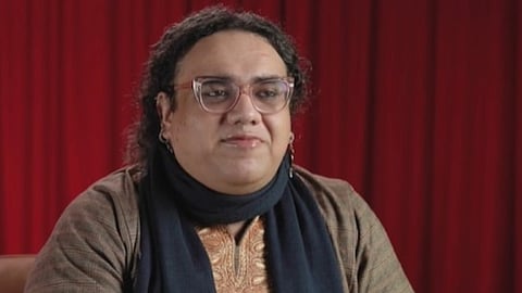 लाल बैकग्राउंड के सामने चश्मा पहने ट्रांसजेंडर एक्टिविस्ट, जो पाकिस्तान के 'मर्दानगी' दावों को चुनौती देने और लंदन में शरणार्थी बनने के संघर्ष के बावजूद सकारात्मक मुद्रा में दिख रही हैं
