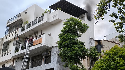 ऊपरी मंजिल से घना काला धुआँ निकलती एक बहुमंजिला इमारत, जहाँ बालकनी में लोग खड़े हैं और बचाव के लिए सीढ़ी लगाई गई है।