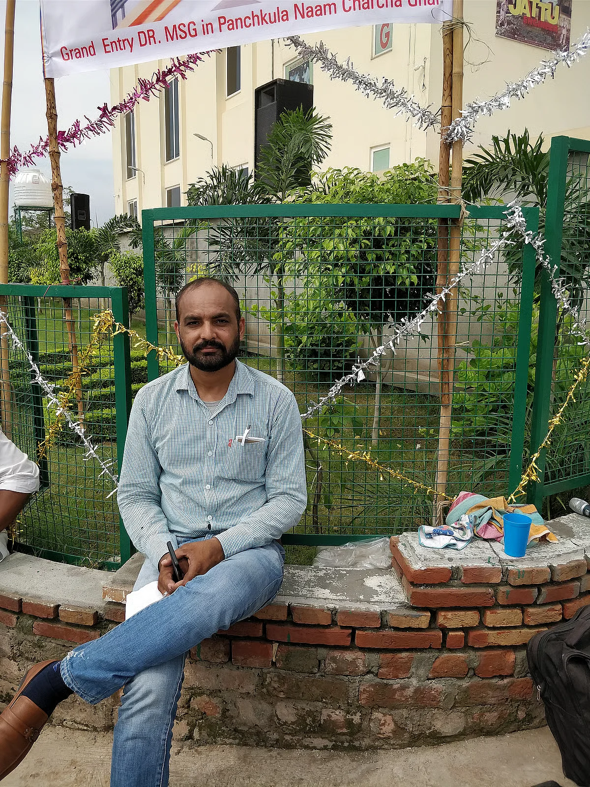 Vajinder Insaan.  (Picture of a man sitting on a low brick wall with greenery and a building behind him.)