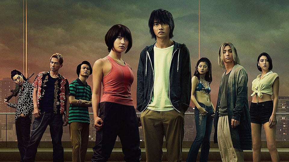 An ensemble cast photo from the Netflix live-action series Alice in Borderland, featuring Arisu, Usagi, and other key survivors standing on a rooftop against a dystopian, sunset-lit Tokyo skyline.