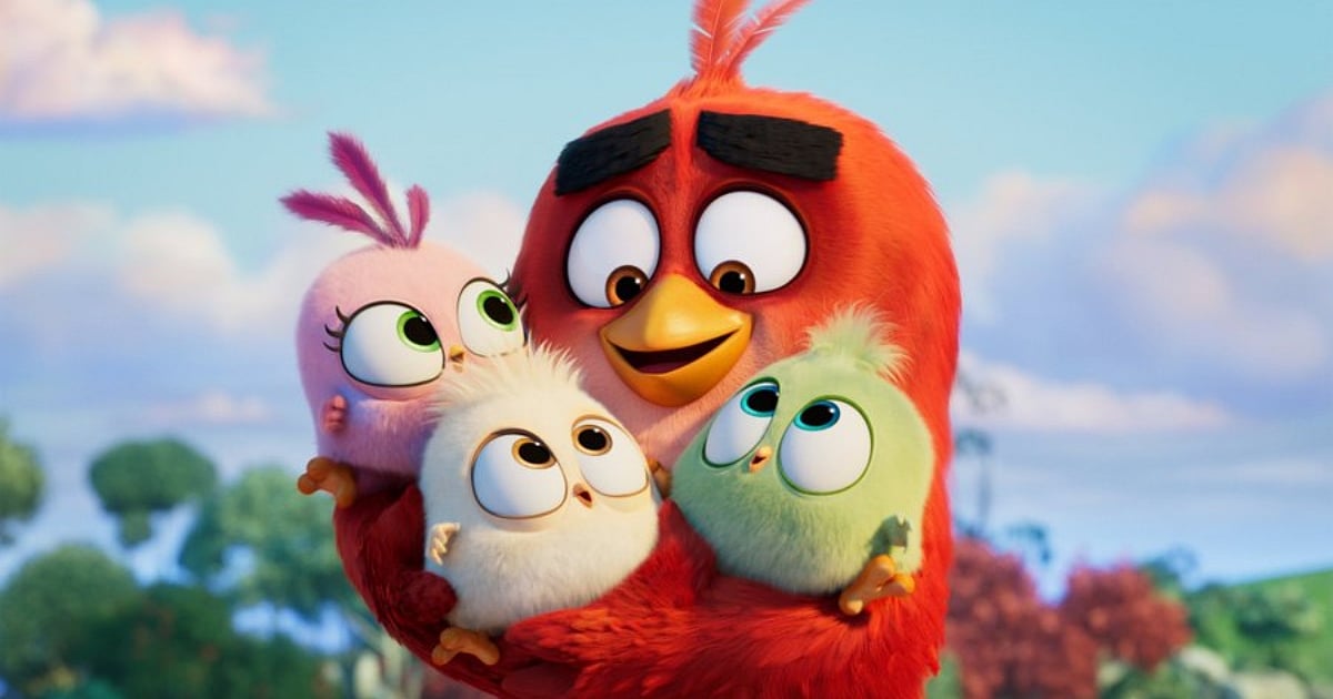 A heartwarming close-up from The Angry Birds Movie showing Red, the large red bird, smiling and hugging three adorable, round hatchlings in pink, white, and light green colors against a soft, outdoor background.