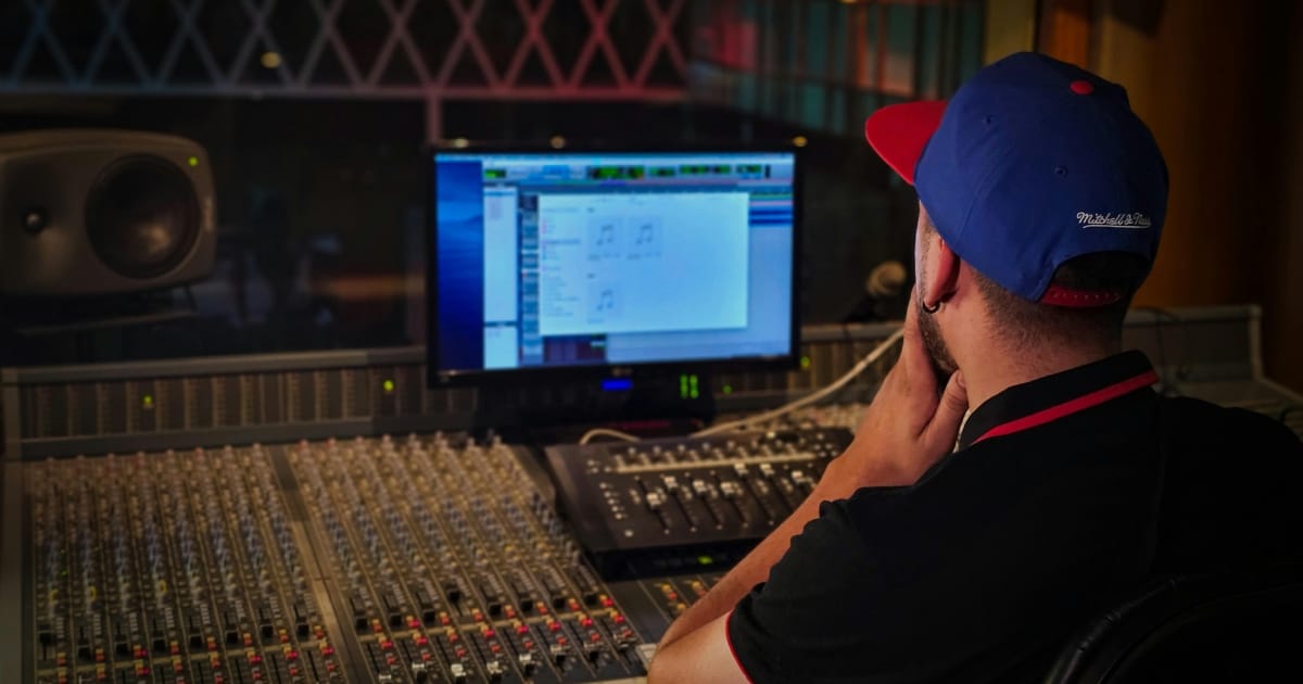 Rear view of a man wearing a blue and red baseball cap sitting in front of a large professional audio mixing console in a recording studio, looking at a computer screen displaying audio software.