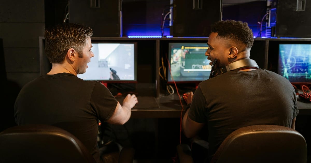 Rear view of two men sitting at a desk in a dimly lit room, smiling and talking while playing video games on separate monitors.