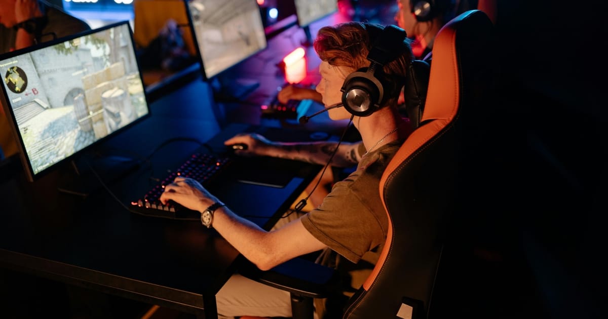  A young man with red hair wearing a headset and a mechanical watch, intensely focused while playing a first-person shooter game on a large computer monitor in a dark gaming room with neon lights.
