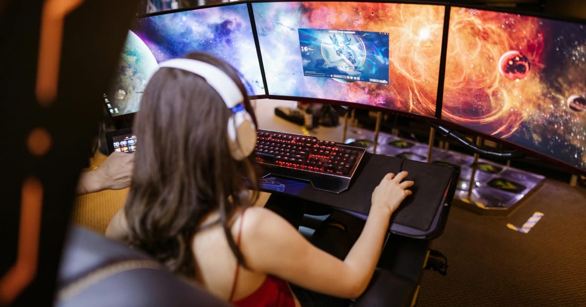  Rear view of a woman with long brown hair wearing white headphones, playing a video game across a massive three-monitor panoramic setup displaying a vibrant space nebula.