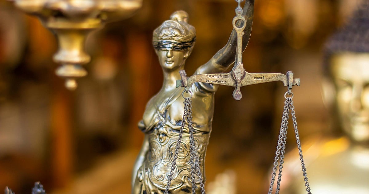 A shallow depth-of-field close-up of a bronze or brass Lady Justice statue holding balanced scales. The figurine wears a blindfold and an ornate, flowing robe with intricate engraved patterns across the torso. 