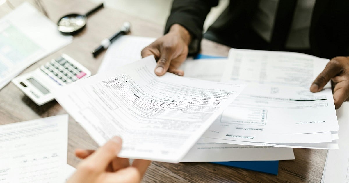 A close-up photo of two people exchanging tax paperwork across a wooden desk in an office setting. One person in a dark business suit holds a stack of printed documents while another person reaches forward to receive or review them
