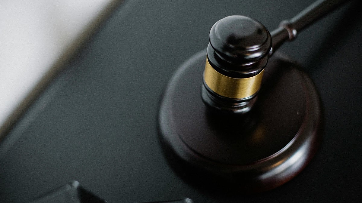 A close-up image of a dark wooden judge’s gavel resting on its circular sound block atop a smooth black surface. The gavel features a polished black finish with a metallic gold band around the head, reflecting soft ambient light. 