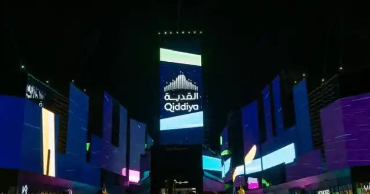 A large, dark structure featuring a prominent, glowing vertical screen displaying the "Qiddiya" logo in both Arabic and English. The surrounding architecture is also illuminated with vibrant blue and purple neon lights against the night sky.