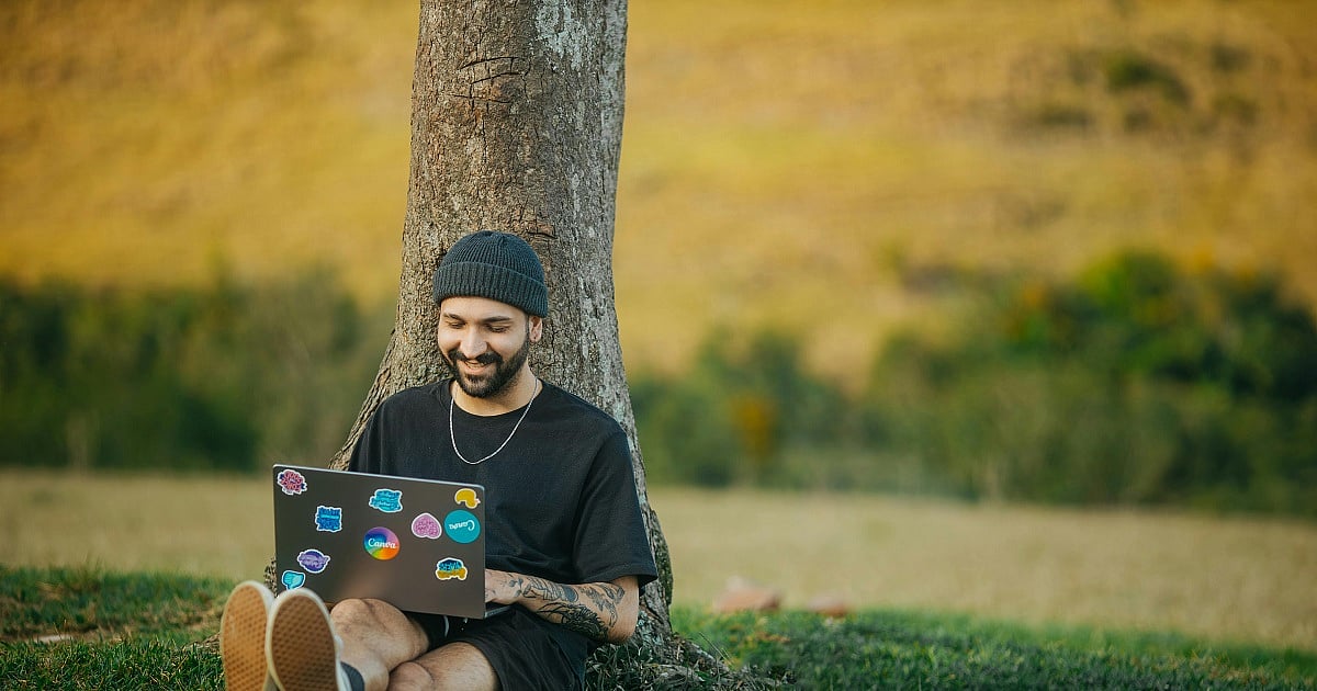 A person sits beneath a large tree in a rural outdoor setting, using a laptop decorated with colorful stickers. Wearing a dark beanie and black T-shirt, they smile while working remotely on the grass. The blurred countryside backdrop features open fields and natural greenery, illustrating how rural broadband and Wi-Fi connectivity can enable remote work, online learning, and digital access outside urban areas.