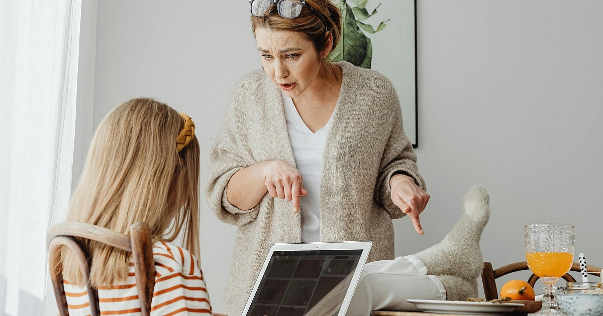 A tense interaction unfolds at a dining table as an adult woman appears to scold or reprimand a young girl seated across from her with a laptop. The woman, wearing a light beige cardigan over a white shirt, leans forward with a stern expression and points both index fingers downward while speaking.