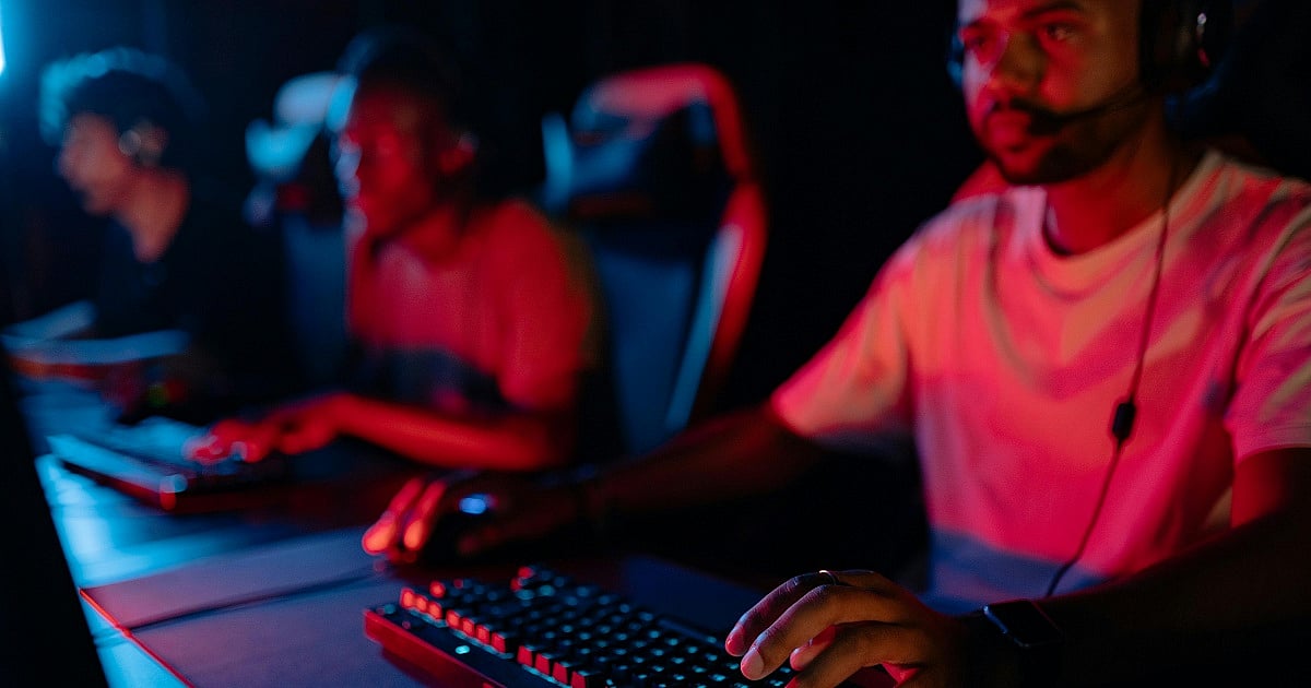 Three gamers sit side by side at desktop gaming stations in a dimly lit cyber café. The foreground focuses on a player wearing a white T-shirt and a gaming headset with a microphone, resting one hand on a mouse and the other near a mechanical keyboard illuminated by blue and red lighting.