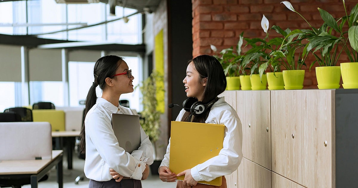 Two women stand and chat in a modern open-plan office beside a row of light wood storage cabinets topped with bright green potted plants. The woman on the left wears glasses with red frames, a white blouse, and dark trousers, holding a closed laptop against her chest while listening attentively. The woman on the right wears a white blouse and carries a large yellow folder, with a headset resting around her neck as she speaks.