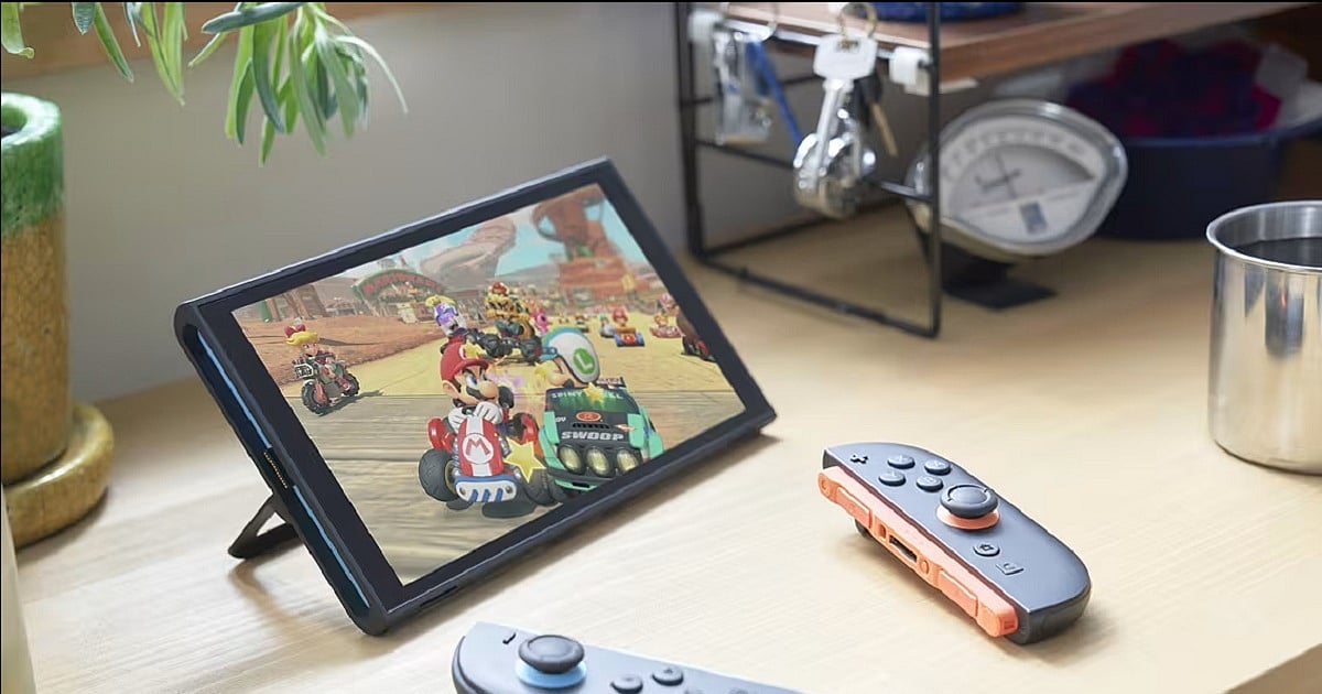 Promotional image showing a Nintendo Switch in tabletop mode resting on a light wooden desk. The screen displays a Mario Kart race featuring Mario and several other racers speeding through a desert-themed track. Two detached Joy-Con controllers lie on the desk in the foreground. Surrounding the console are household items including a potted plant, a metal cup, a set of hanging keys, and a vintage-style analog scale on a shelf in the background.