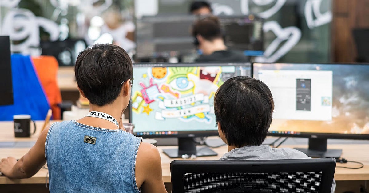 Two Ubisoft employees sit side by side at a shared workstation in a modern open-plan office, viewed from behind as they work on multiple monitors. One screen displays colorful concept art and graphics, while another shows design software. Blurred coworkers and decorative Ubisoft-themed window decals appear in the background of the studio.