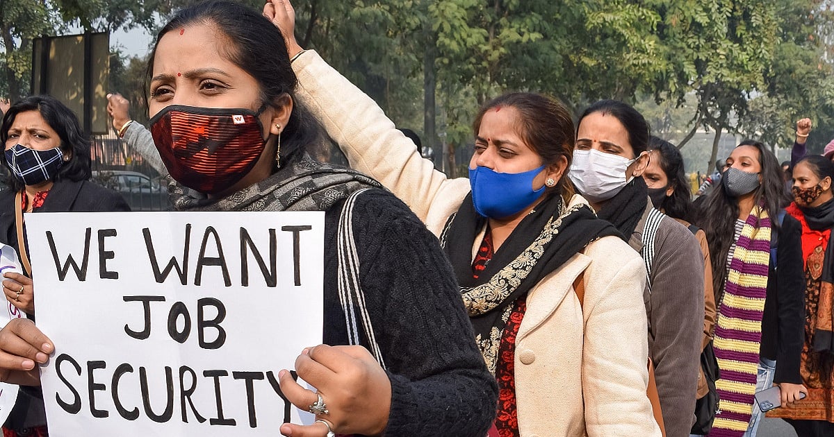 A group of women wearing face masks participate in a protest march in India, gathered outdoors on a tree-lined street. In the foreground, one woman holds a handwritten placard reading “WE WANT JOB SECURITY,” while others behind her raise their hands and chant slogans. The scene reflects a demonstration advocating for workers’ rights and employment security.