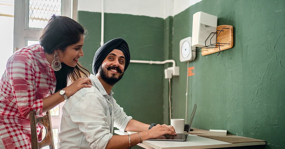 A man wearing a black turban sits at a small desk working on a laptop while smiling toward a woman leaning over his shoulder. The woman, dressed in a pink-and-white checkered top with large earrings, rests a hand on his shoulder. The room features green walls, a window letting in natural light, exposed wiring, and minimalist wall fixtures.