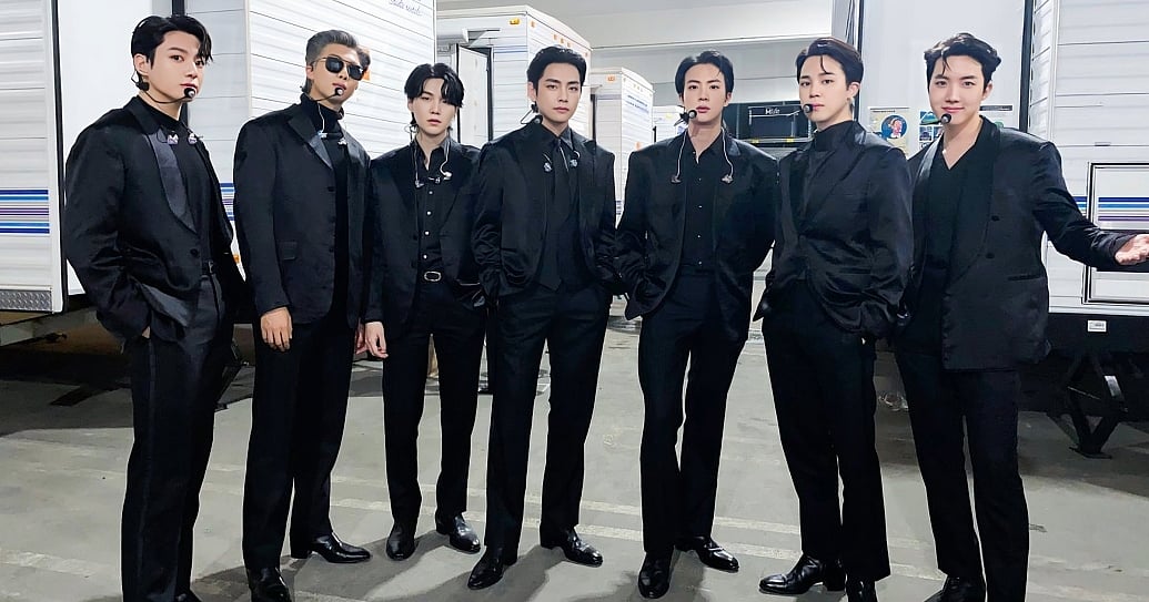 The seven members of the K-pop group BTS stand side-by-side in a row, posing confidently backstage. They are all dressed in sleek, matching all-black formal suits, button-down shirts, and dress shoes, with member RM also wearing sunglasses. They are equipped with headset microphones and in-ear monitors, standing on a concrete lot with white production trailers lined up in the background.