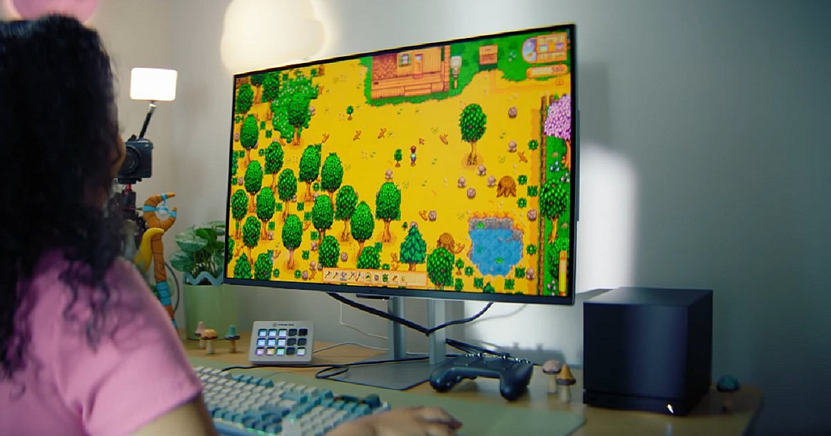 A person plays Stardew Valley on a large monitor using a Steam Deck connected in docked mode. The gaming setup includes a mounted light, small desk decorations, a streaming deck device, and a black Steam Controller.