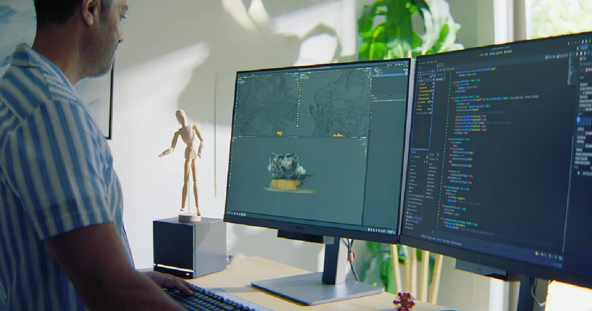 A game developer works at a dual-monitor setup, with one screen displaying a 3D scene in a game engine and the other showing code. A Steam Deck sits on the desk below the monitors, partially visible beneath the screens. The room is brightly lit with plans creating a vibrant atmosphere.