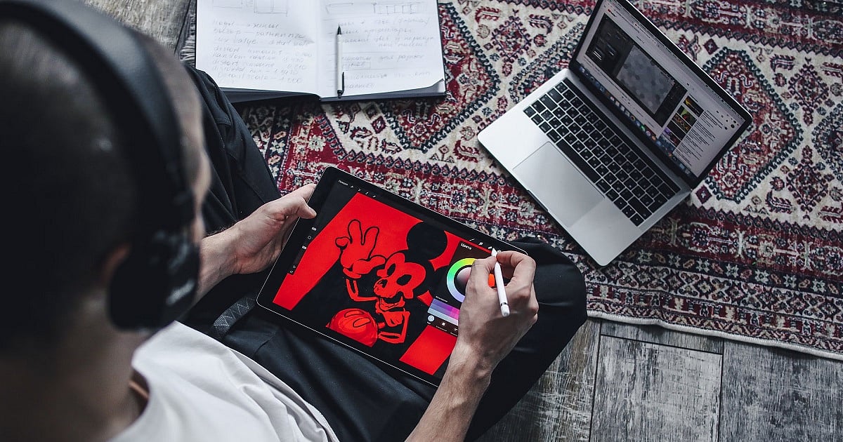 An animator wearing headphones works on a digital illustration using a stylus and tablet while seated on the floor. The tablet displays a bold red-and-black cartoon character, with a laptop showing reference material and an open notebook nearby, creating a home digital art and animation workspace.