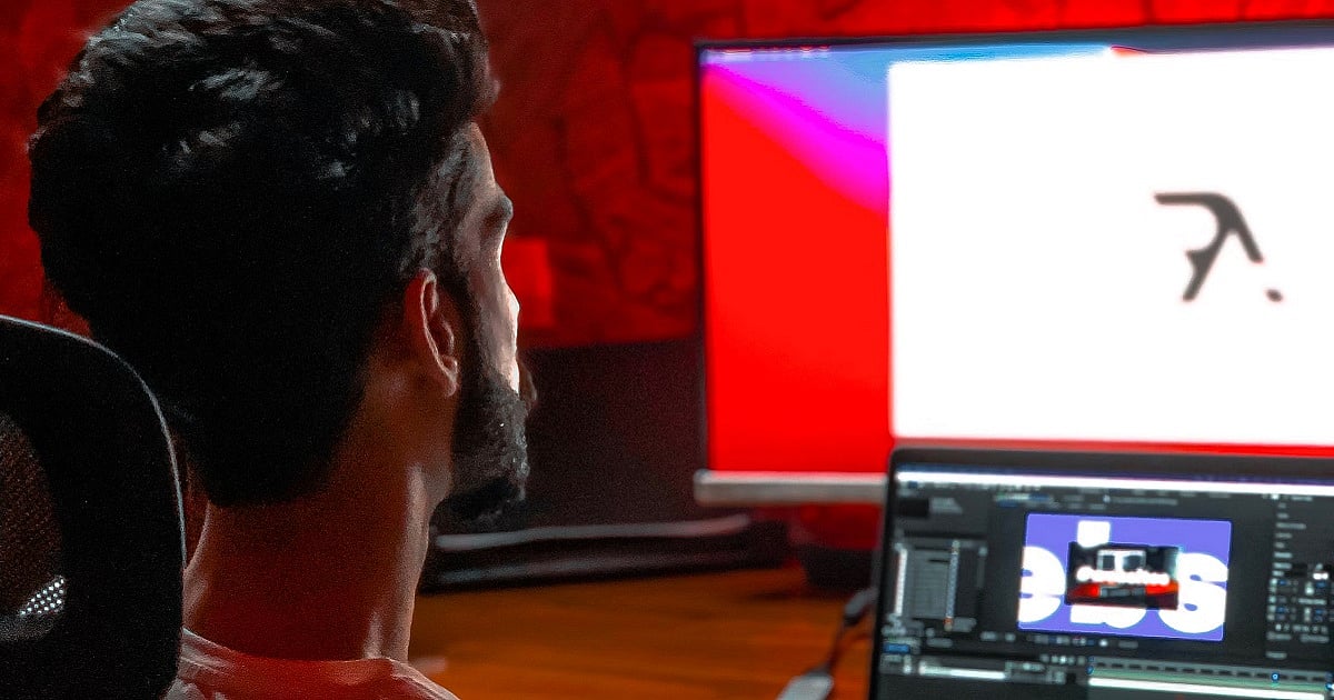A digital animator sits at a desk facing dual monitors in a dimly lit workspace illuminated by red ambient lighting. The larger display shows a minimalist animation frame on a white background, while the laptop below is open to animation software with a project timeline and composition window.