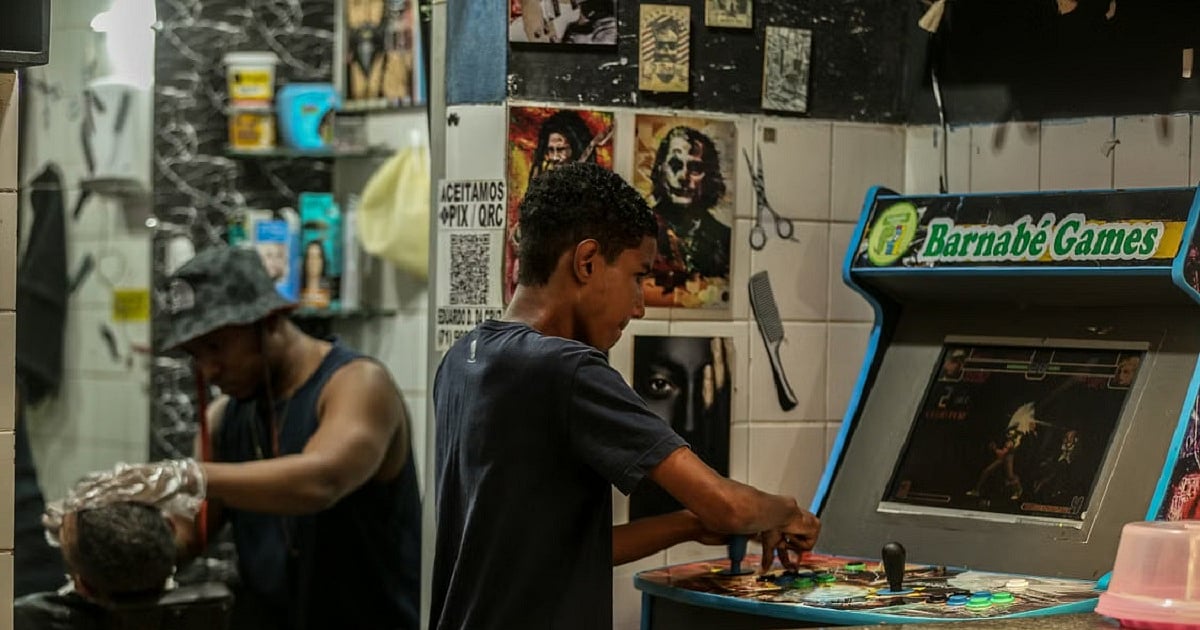 A young man stands at a retro arcade cabinet inside a neighborhood barbershop, focused on a fighting game while gripping the joystick and colorful buttons. In the background, a barber cuts a customer's hair, with mirrors, posters, and grooming tools lining the walls, creating a lively community atmosphere where gaming and everyday life intersect.