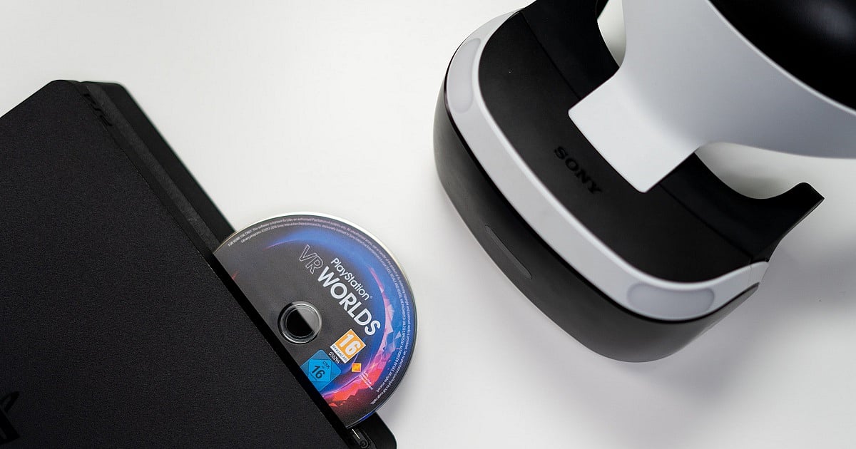 A black PlayStation 4 console ejects a PlayStation VR Worlds game disc while a white-and-black PlayStation VR headset rests nearby on a clean white surface. The composition highlights Sony's first-generation virtual reality hardware alongside the physical game disc.